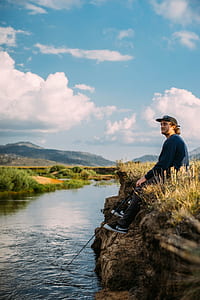 man wearing grey sweatshirt sitting down on grass while fishing near body of water during daytime thumbnail