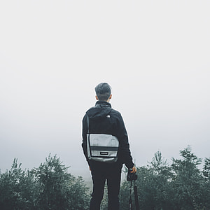 man wearing black hooded jacket and black pants outfit with gray crossbody bag holding black camera body thumbnail