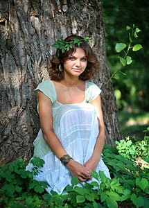 woman wearing teal scoop-neck cap-sleeved dress sitting in front of tree surrounded by green leaf plants thumbnail