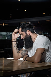 man in white crew-neck t-shirt thinking while leaning on brown wooden table thumbnail