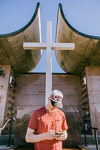 man in orange crew-neck T-shirt using smartphone standing near wooden cross thumbnail