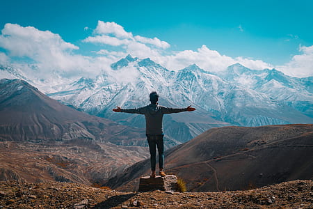 man wearing gray long-sleeved shirt and blue denim jeans standing next to mountain range during daytime thumbnail