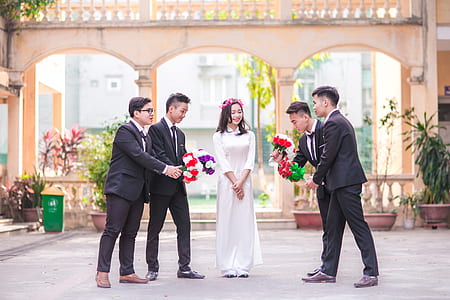 woman wearing white dress standing in the middle of four men offering flowers thumbnail