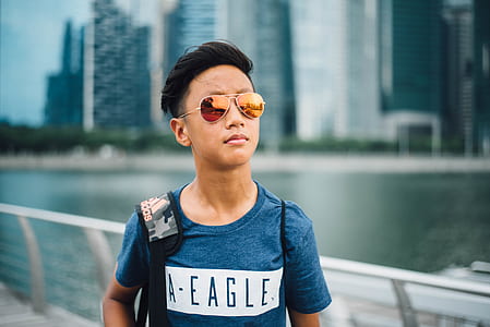boy wearing blue American Eagle crew-neck t-shirt and brown framed Aviator-style sunglasses carrying gray and black camouflage adidas bag thumbnail