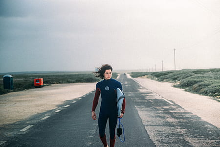 person carrying surfboard while standing on asphalt road thumbnail