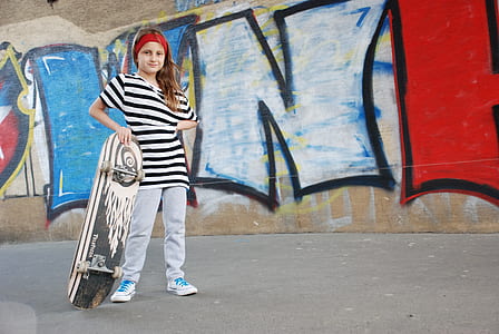 girl holding white and black skateboard standing near gray wall thumbnail