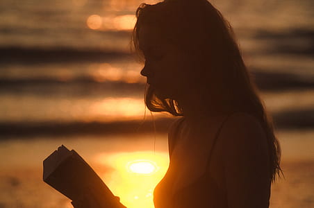 woman reading book near body of water during sunset thumbnail