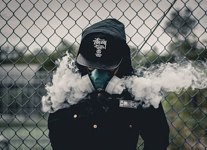 shallow focus photography of person wearing green and white bong mask, black cap, and hooded jacket leaning on gray steel cyclone fence thumbnail