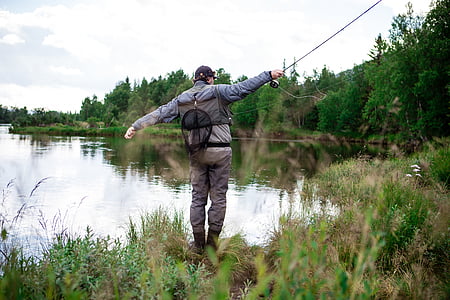 man wearing blue holding fishing rod standing on green grass across body of water outdoors thumbnail