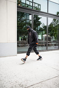 person wearing black capris, hoodie, and balaclava walking by glass and white concrete wall thumbnail