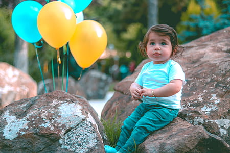 Toddler Wearing White Shirt Sitting on Rock Beside Yellow and Blue Balloons thumbnail