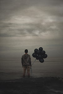 man in gray hoodie standing on body of water while holding black balloons thumbnail