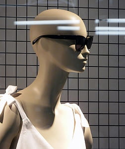 The bald head of a female mannequin wearing a pair of dark sunglasses against a background of strobe lighting reflecting in a shop window thumbnail