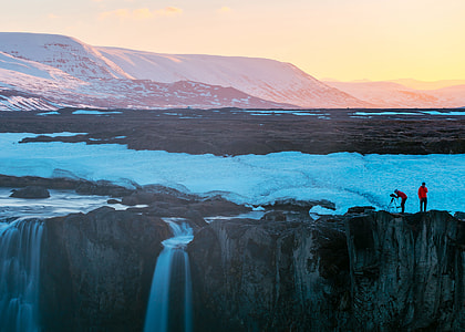 photo of two person taking photo of snow covered mountain with waterfalls during daytime thumbnail