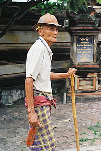 elderly man wearing white polo shirt and orange hardhat walking with brown wooden stick thumbnail
