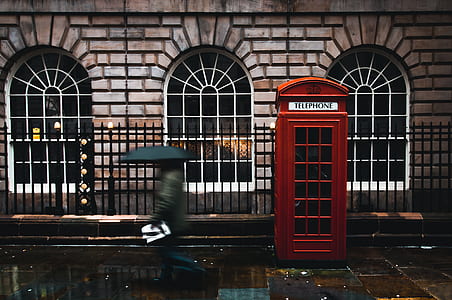 person walking beside red telephone book in front of black metal gate and brown brick building thumbnail