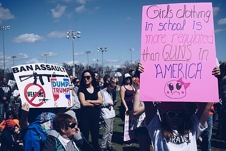 standing woman wearing black crew-neck T-shirt and black sunglasses surrounded by people holding posters under clear blue sky thumbnail