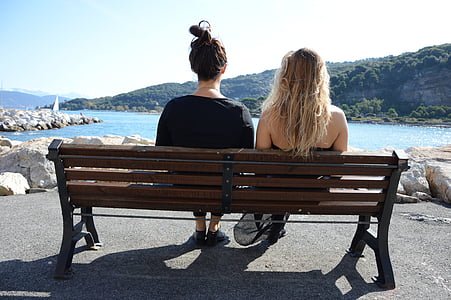 two women sitting on bench near body of water and mountains during daytime thumbnail