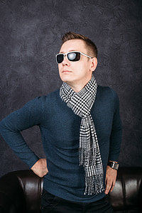 man wearing black sweater and white and black scarf posing in front of black leather sofa thumbnail