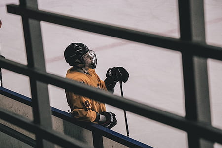 ice hockey player wearing orange jersey shirt and black helmet leaning on gray and blue metal railing thumbnail