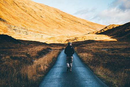 man wearing jacket walking at the road toward brown hill thumbnail