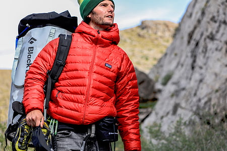 man wearing red bubble jacket and gray backpack standing beside rock thumbnail