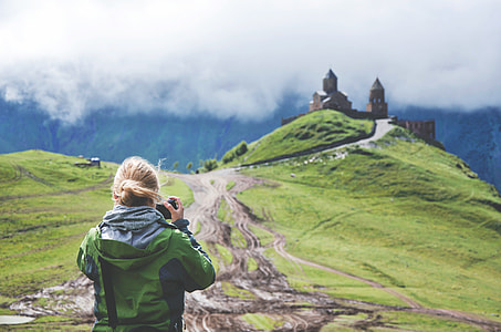 woman in green hooded windbreaker jacket holding dslr camera taking a photo of concrete building under white clouds during daytime thumbnail