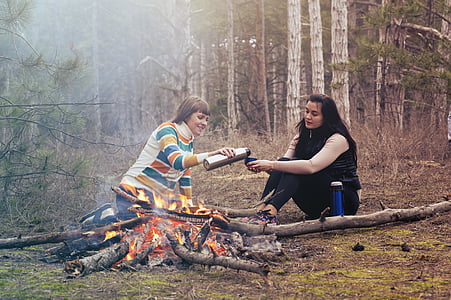woman holding vacuum flask pouring liquid on cup on woman sitting on soil near firepit thumbnail