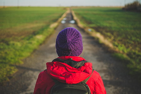 person wearing purple knit cap and red hooded jacket facing concrete road between green grass field during daytime thumbnail