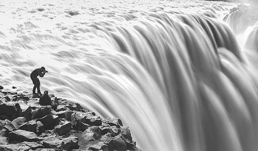 grayscale photo of man talking photo near cliff of waterfall thumbnail