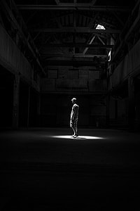 grayscale photo of person standing in middle of empty building with light passing through its roof thumbnail