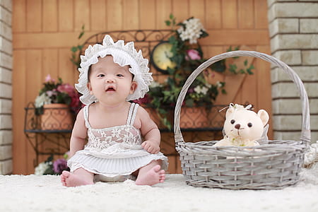 baby wearing white floral dress sitting beside gray basket with white teddy bear inside on fleece mat thumbnail