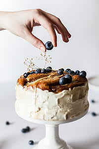 person holding blueberry above cake with white icing thumbnail