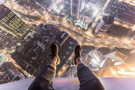 A man sitting on the roof of a city building with his feet and shoes hanging over the edge thumbnail