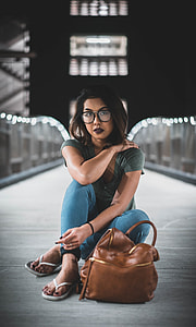 woman in gray top and blue jeans sitting on pathway beside brown leather bag during daytime lowlight photography thumbnail