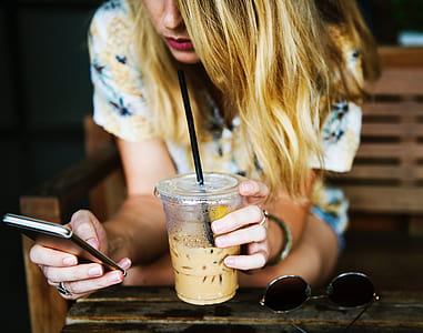 woman wearing white and multicolored floral shirt sitting on brown bench holding disposable cup and using smartphone thumbnail