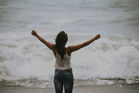 woman wearing white spaghetti strap top and blue bottoms raising hands facing ocean waves thumbnail