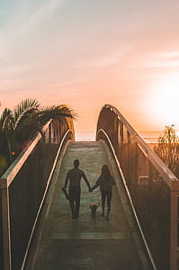 man and woman walking on brown wooden bridge near coconut tree during sunset thumbnail