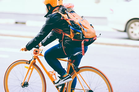 selective focus photography of person wearing black jacket, orange backpack, blue denim jeans, and black-and-white Puma sneakers riding orange bicycle on street thumbnail