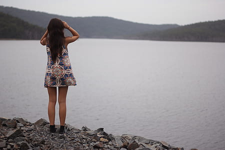 woman standing on big rock beside body of water under gray sky at daytime thumbnail