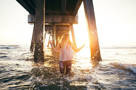 woman wearing white shirt standing on blue body of water under bridge during daytime thumbnail