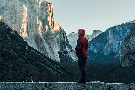 man in hooded jacket standing on concrete floor while looking on gray and brown mountains during daytime thumbnail