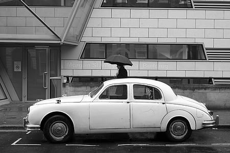 white sedan parked beside building with man walking white holding umbrella thumbnail