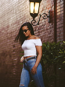 woman wears white off-shoulder crop top and blue denim jeans beside black outdoor lantern and green plants thumbnail