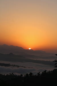 silhouette of trees covered with clouds and mountain in distance during golden hour thumbnail
