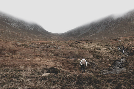 man wearing brown and white top walking on rocky terrain with brown grass thumbnail