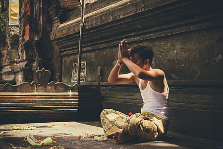 man in white tank top showing praying hand while sitting near brown wall during daytime thumbnail