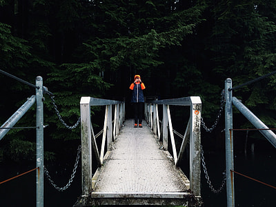person standing on gray footbridge leading to forest thumbnail