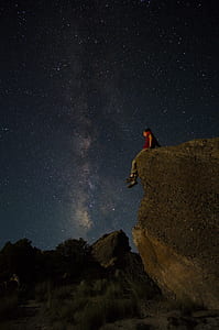 man in red t-shirt sitting on boulder looking at the sky full of stars thumbnail