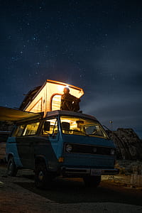 person sitting on top of teal van with camping trailer during nighttime thumbnail